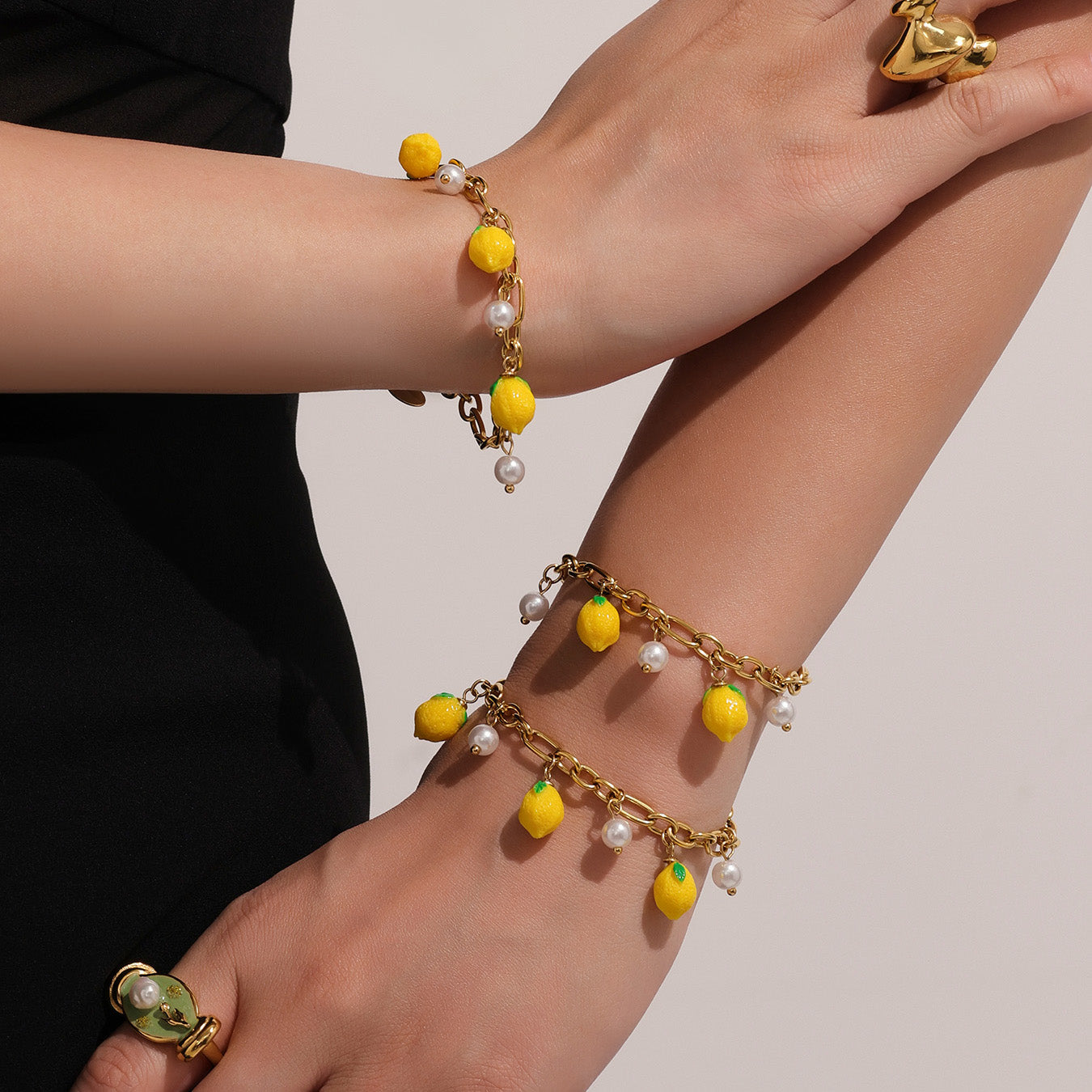 Two wrists wearing gold bracelets with yellow and green lemon charms on a neutral background