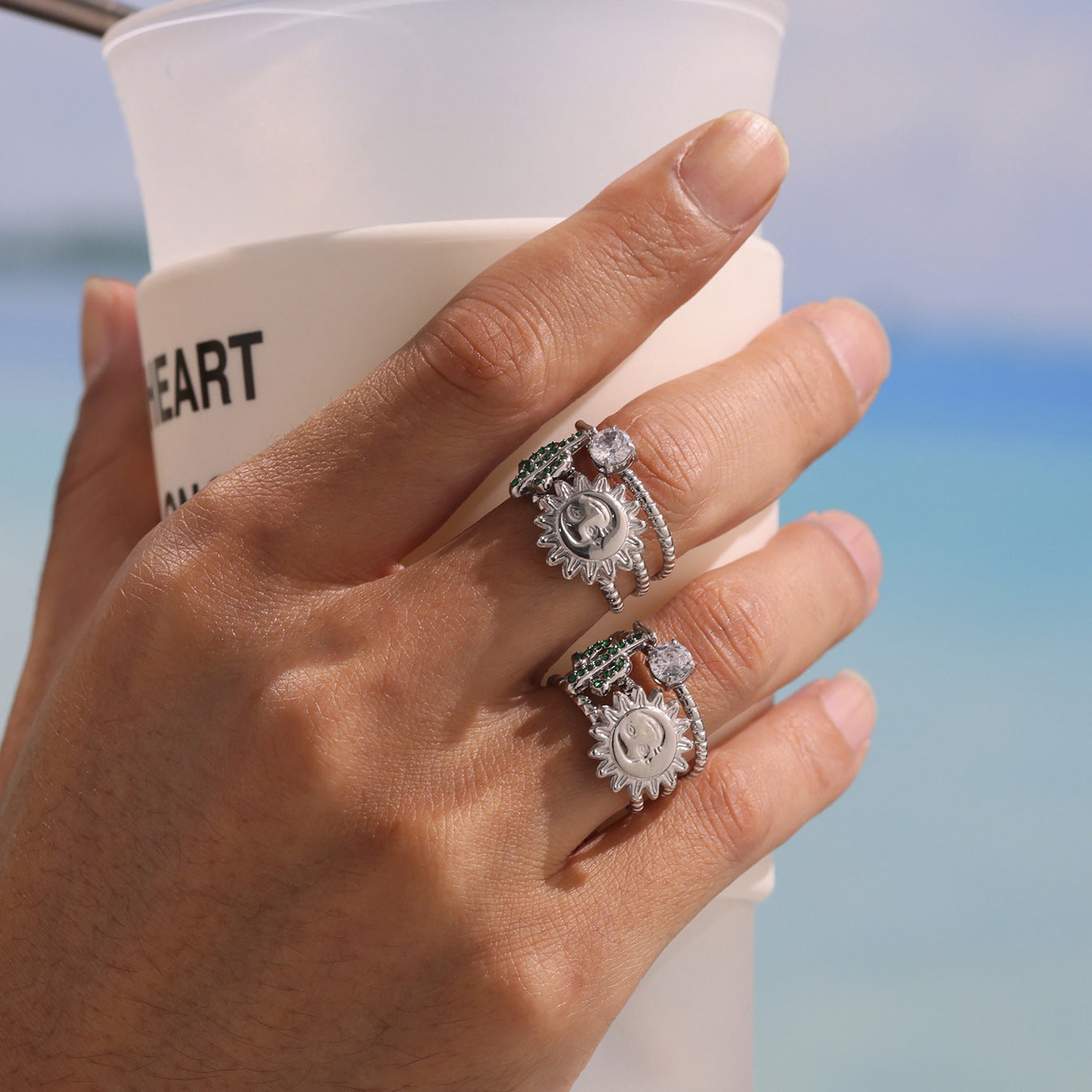 Hand wearing two chunky silver rings with gemstones, holding a white cup against a blurred blue sky background.