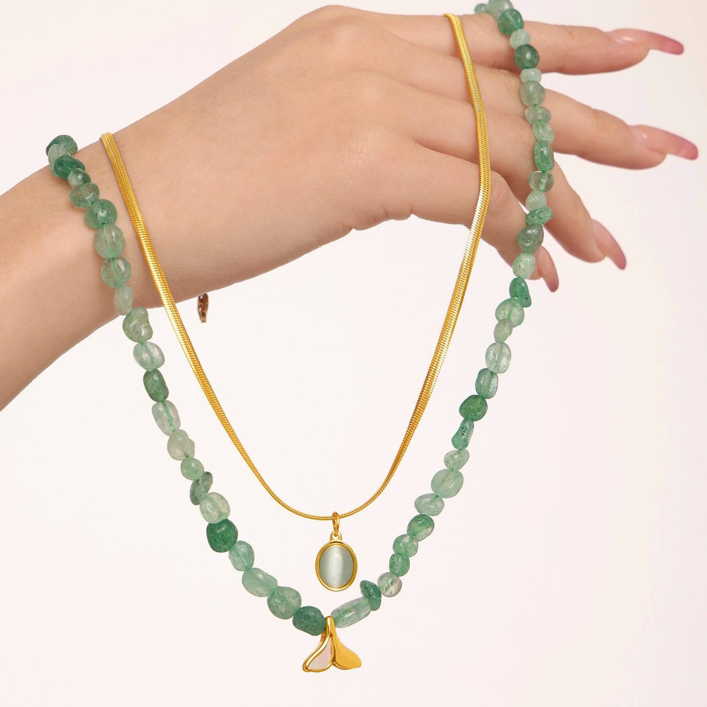 Hand wearing two green beaded necklaces with a gold chain on a light pink background