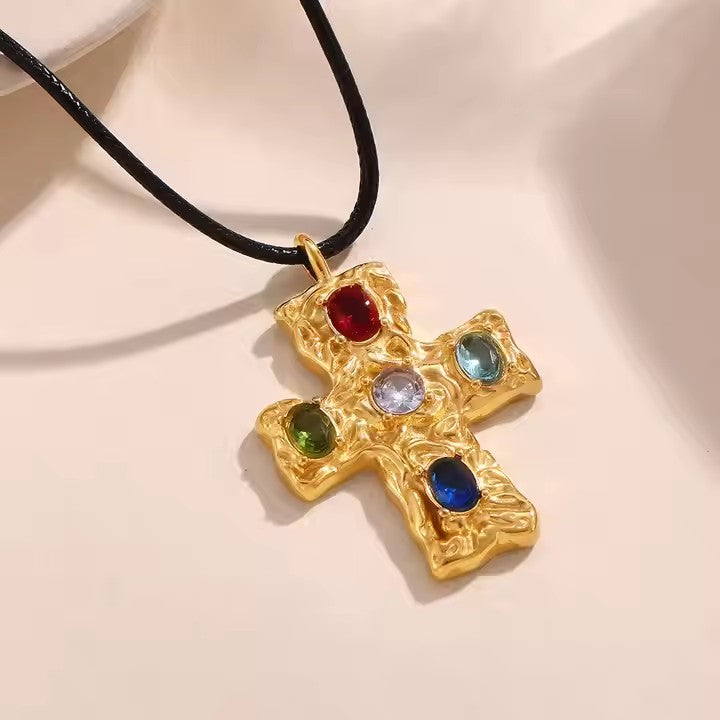 Gold cross pendant with colored stones on a black cord against a beige background