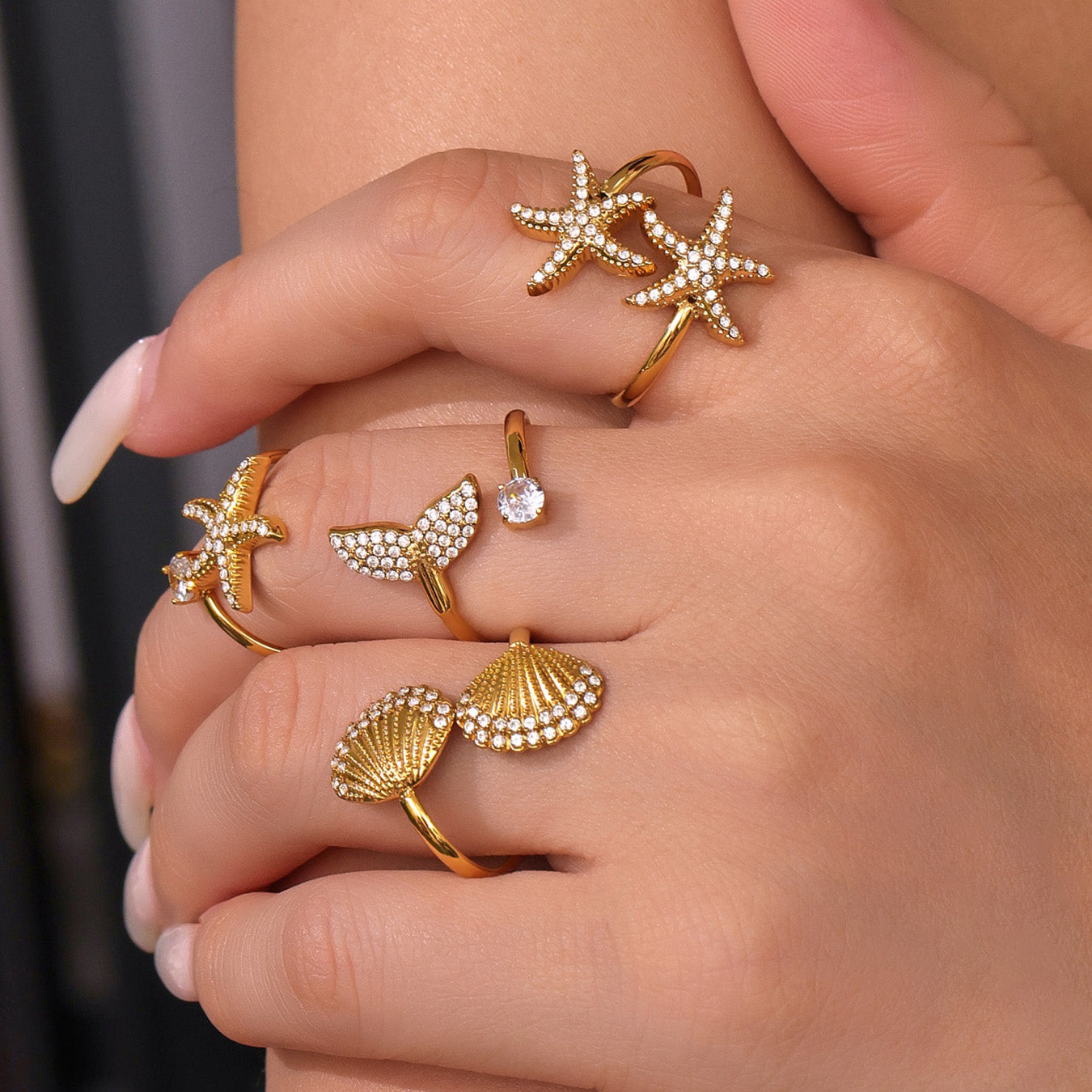 Gold rings with starfish, shell, and diamond designs on a hand against a neutral background