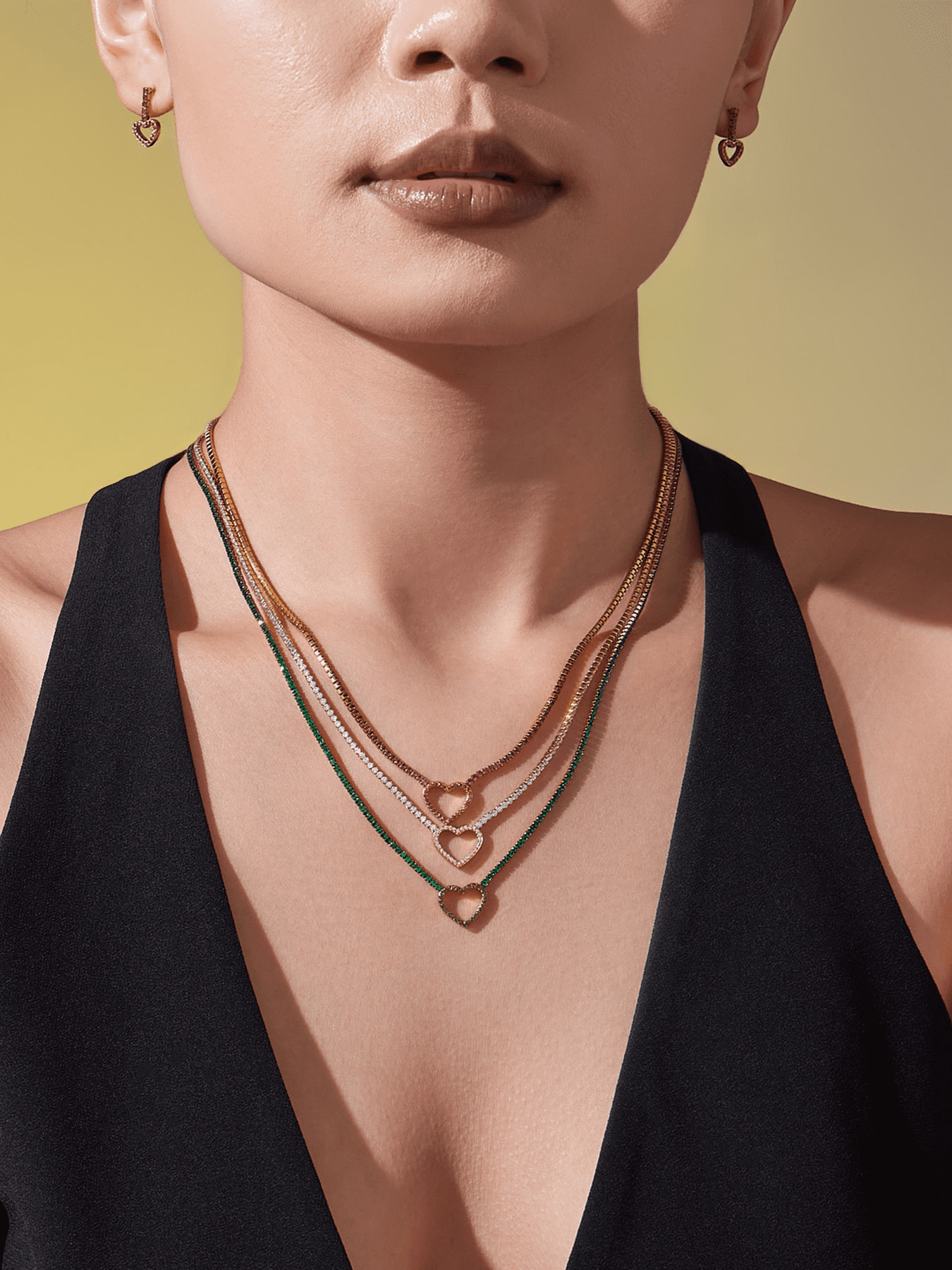 Model wearing layered heart CZ tennis necklaces in gold, green, and red stainless steel chains