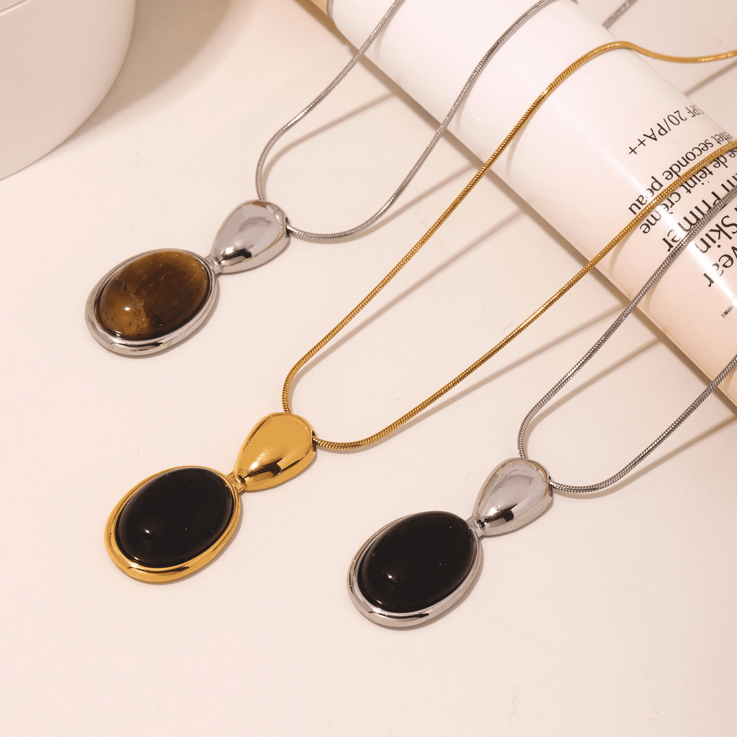 Two lariat necklaces with gold and silver color accents, featuring black oval pendants, displayed against a white background with textural elements.