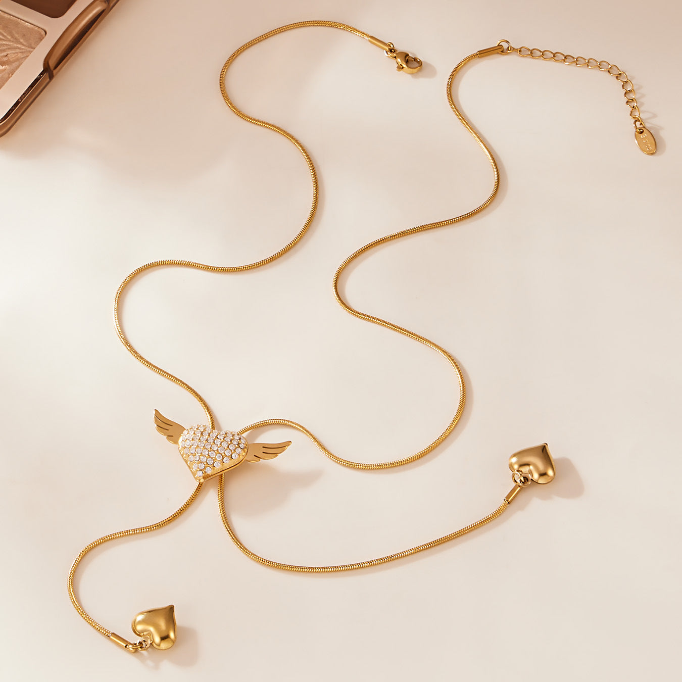 Gold angel wing heart necklace laid flat to show the smooth snake chain, slider detail, and dangling puffy heart charms.