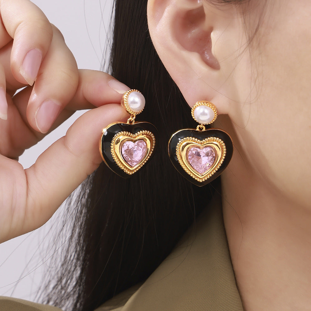 Flat-lay of pink heart pearl stud earrings with black enamel border placed on fabric background, elegant romantic style