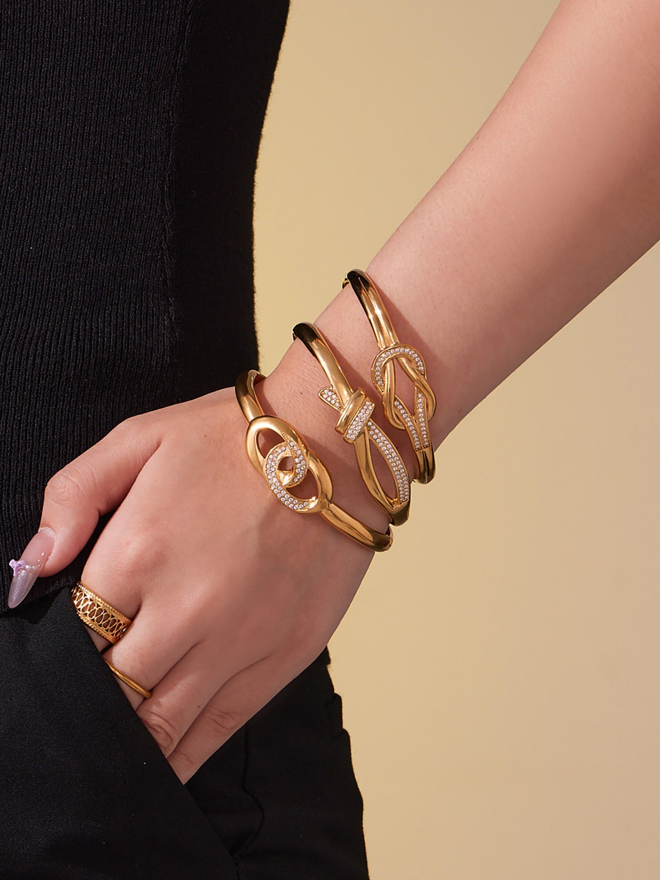 Close-up of a model’s wrist layered with gold knotted CZ cuff bangles, highlighting the pavé zircon shine and smooth stainless steel finish.