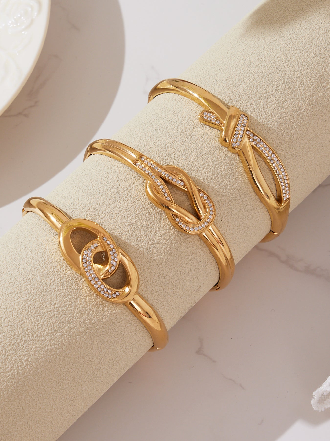 Three gold knotted CZ cuff bangles displayed on a soft textured roll, showing the elegant knot patterns and micro-pavé detailing of each design.
