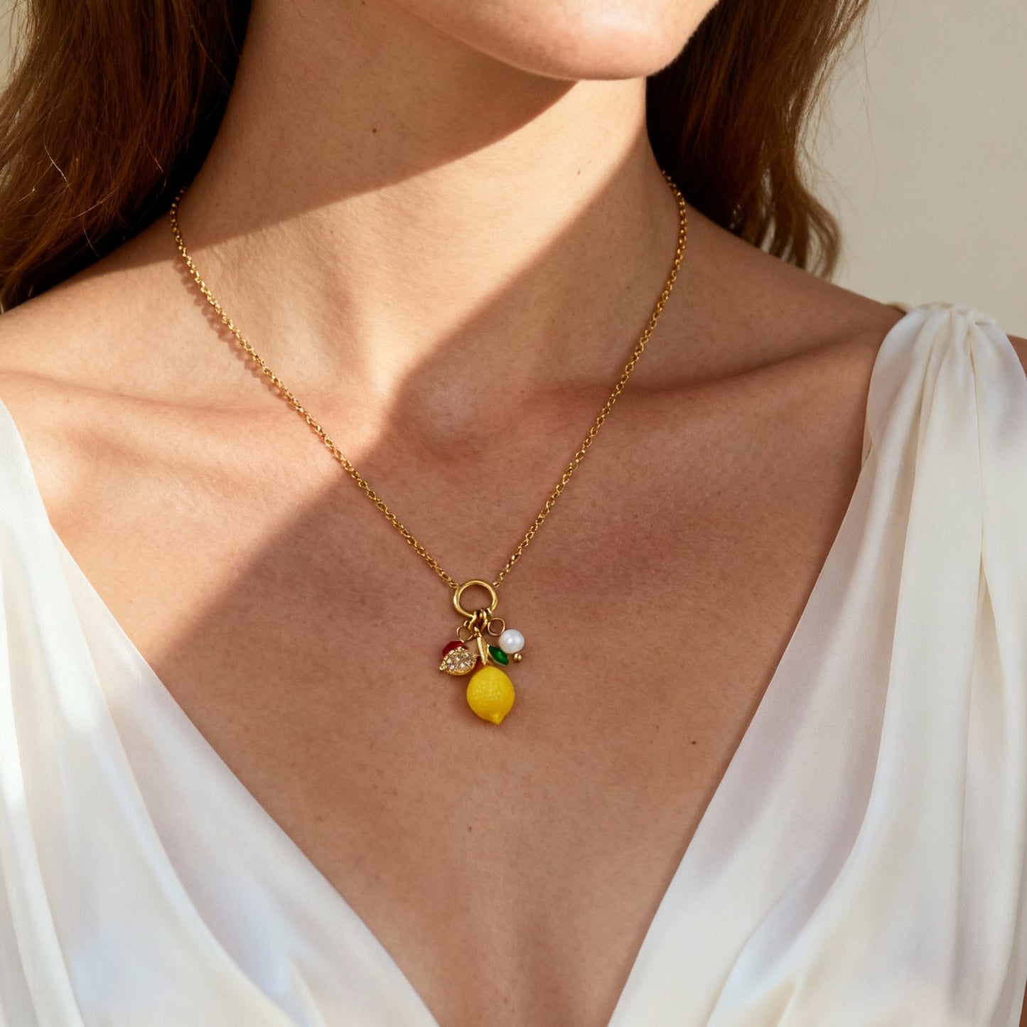 Model wearing the gold lemon charm necklace, featuring a bright yellow resin lemon pendant with red zircon and green leaf accents, shown against a deep V white top in natural light.