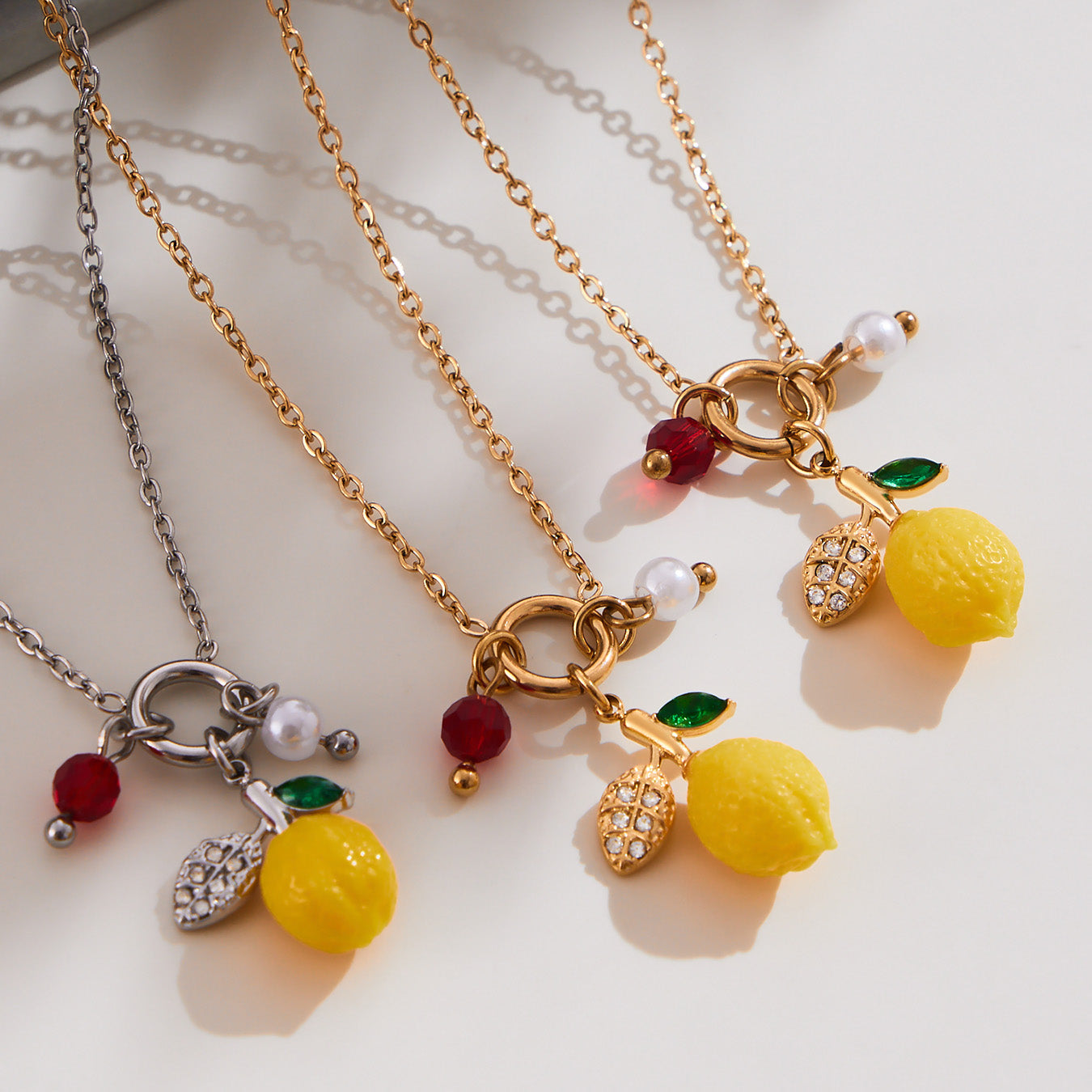 Multiple gold lemon charm necklaces laid out together, each with resin lemon pendants, imitation pearls, red zircon crystals and green leaf charms, highlighting waterproof stainless steel chains