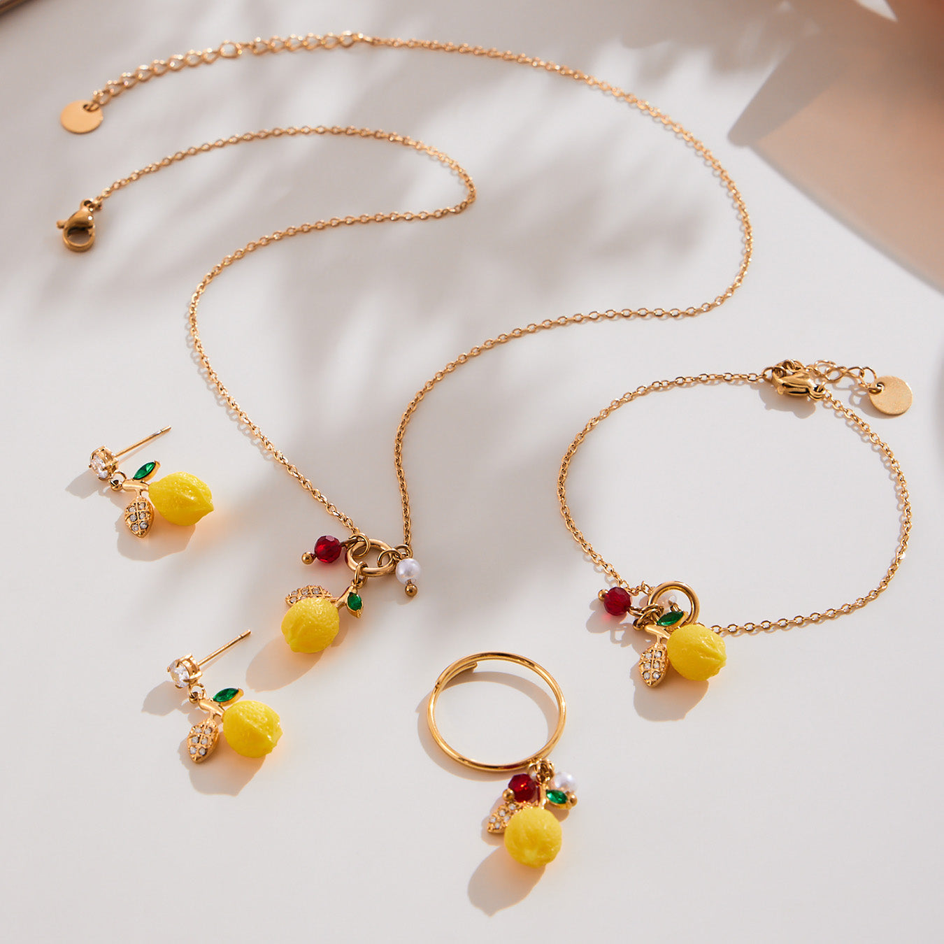 Gold stainless steel lemon charm necklace photographed with matching fruit-inspired earrings, showing resin lemon pendants, red zircon accents, green leaf crystals and imitation pearls arranged together on a soft fabric background.