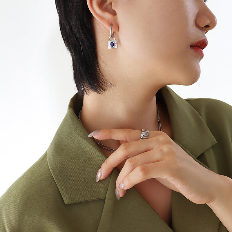 Close-up of gold padlock earrings with green and purple gemstones arranged on a soft neutral background.