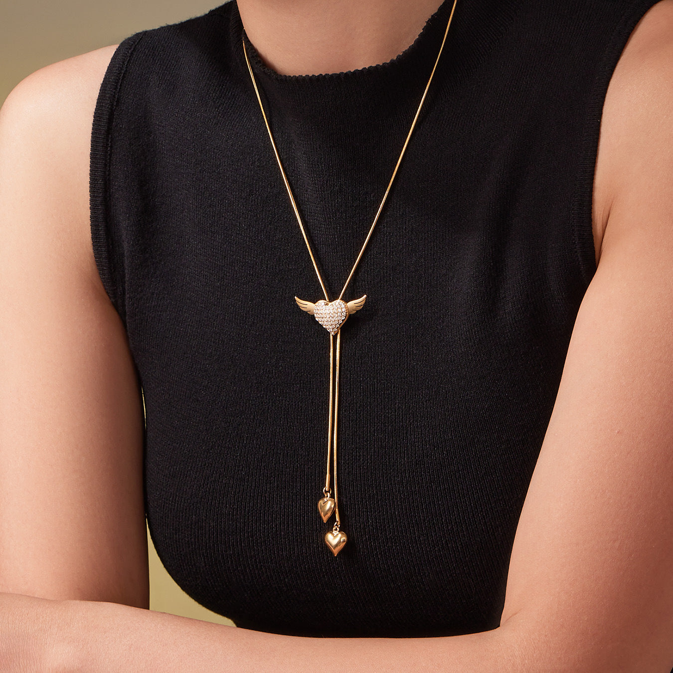 Model wearing the gold winged puffy heart necklace over a black top, showing how the tassel hearts sit at the collarbone.