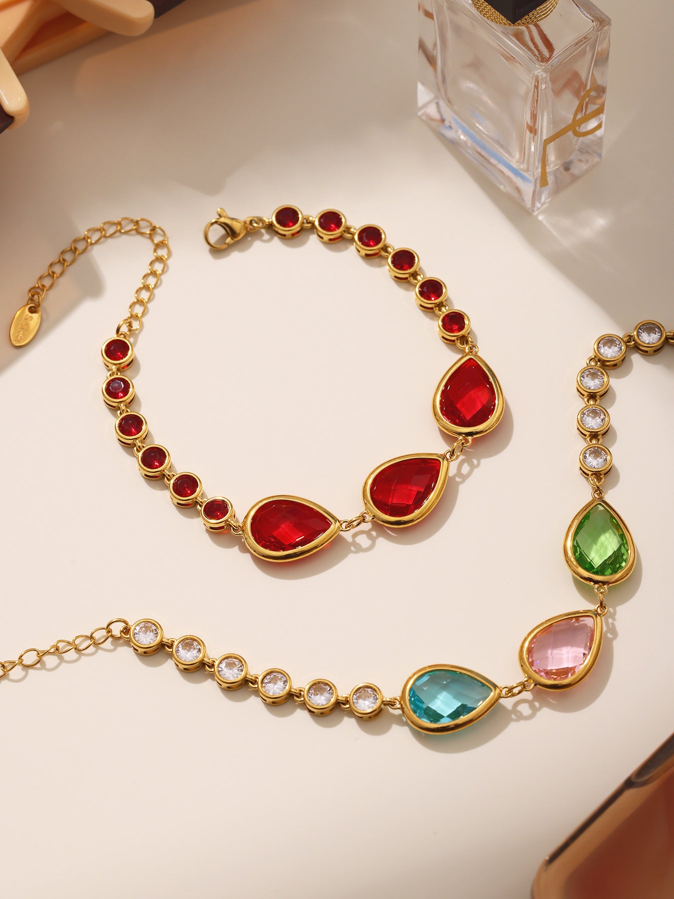 Close-up of red and multicolor teardrop bracelets with round bezel stones, shiny gold stainless steel, lifestyle desk setup