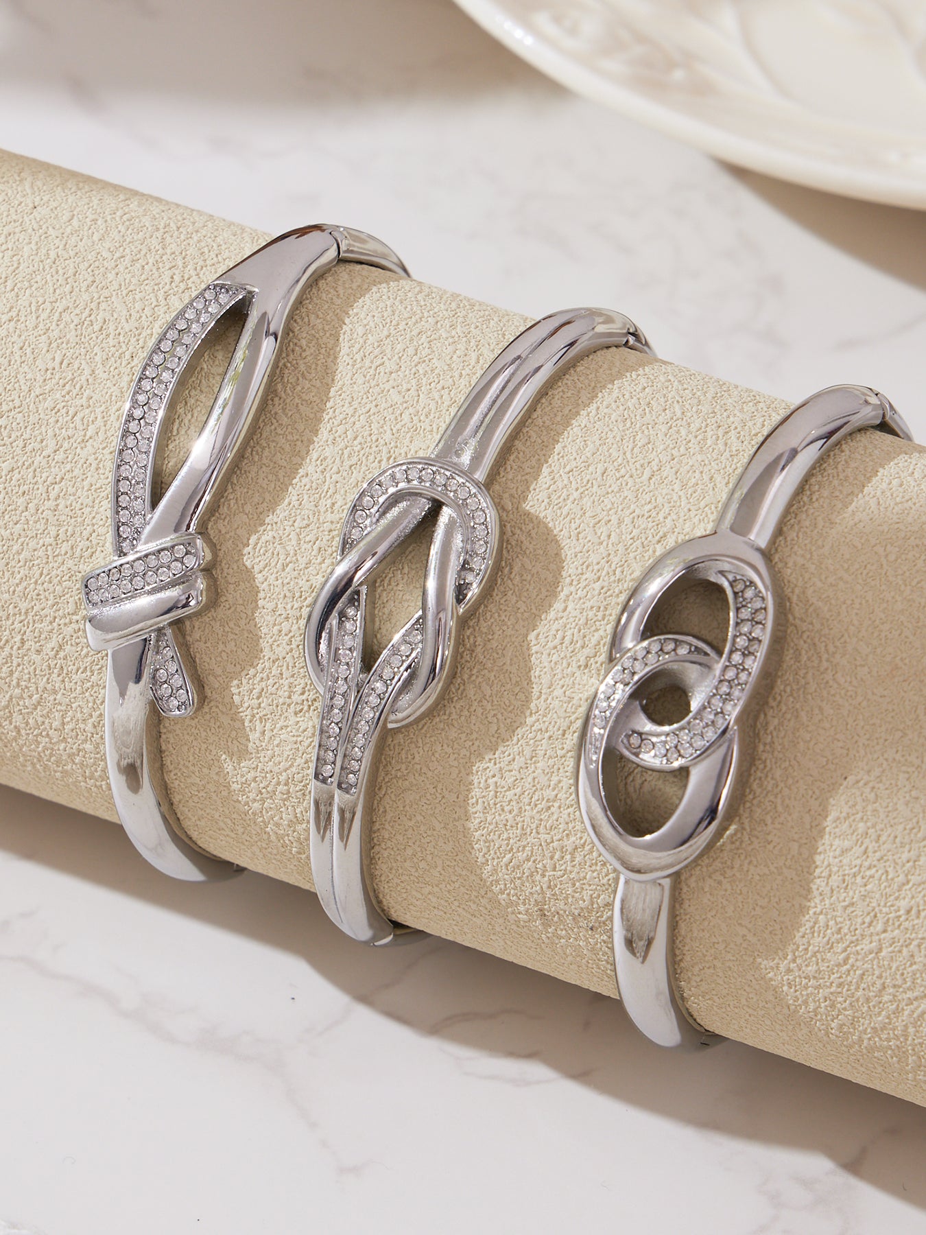 Three silver knotted CZ cuff bangles arranged on a cream display stand, highlighting the reflective stainless steel finish and delicate zircon shine.
