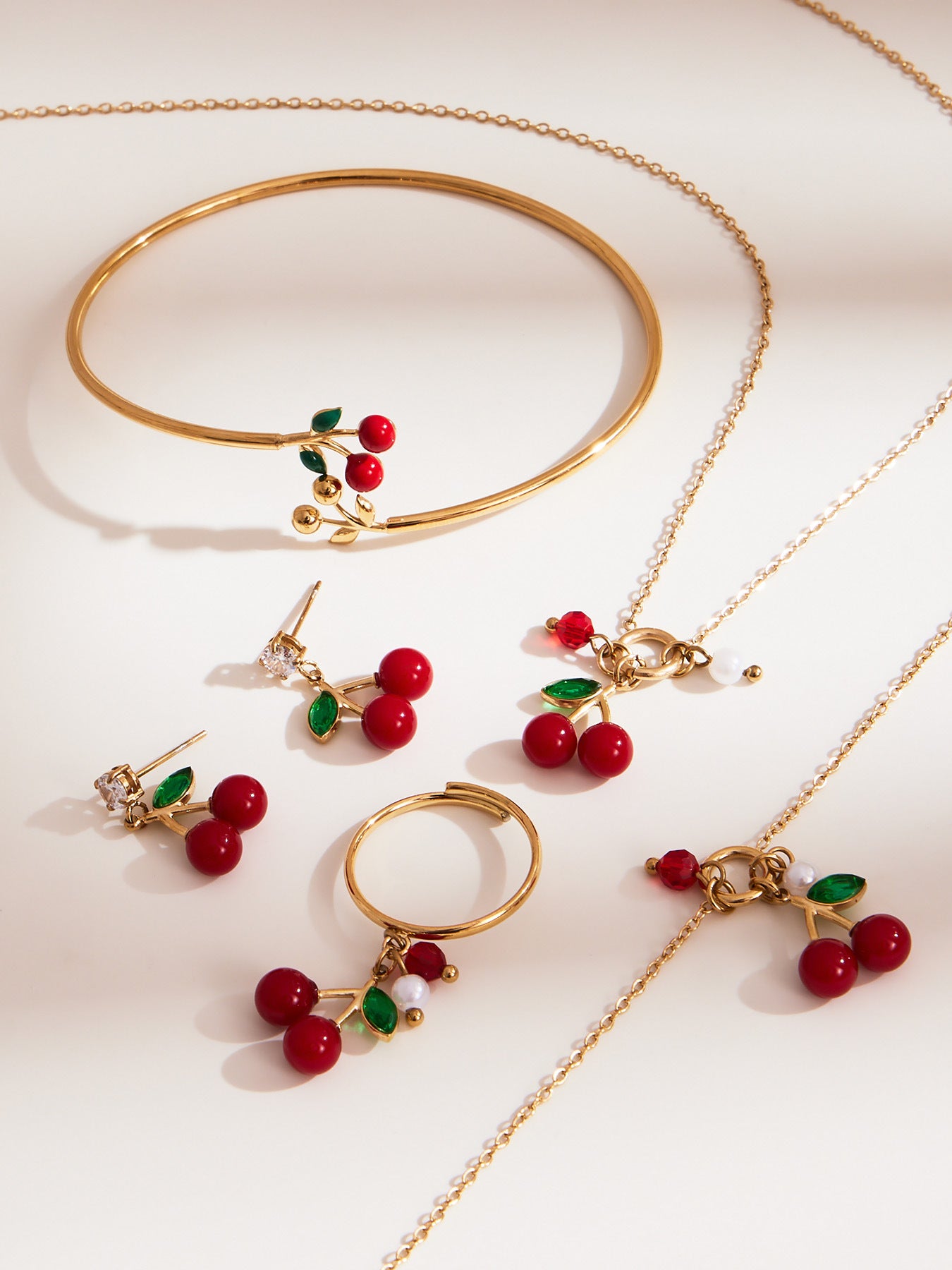 gold cherry jewelry set including cherry necklace, cherry bracelet, cherry studs, and cherry ring, arranged on beige background
